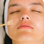 Spa Services - Top view skilled female cosmetologist in uniform and gloves applying essential oil on young woman face skin while working in contemporary spa center