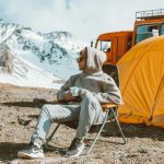 Travel Experience - Full length of male camper sitting on folding chair next to tent in middle of mountain valley and enjoying views