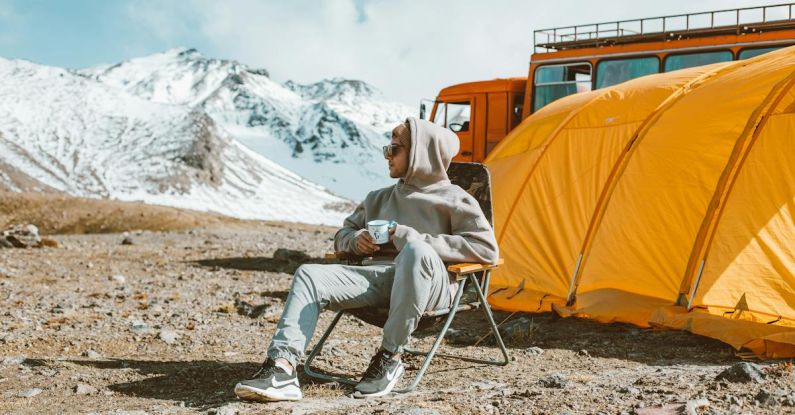 Travel Experience - Full length of male camper sitting on folding chair next to tent in middle of mountain valley and enjoying views