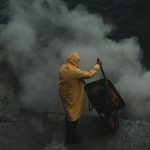 High-Altitude - A man in a yellow rain coat standing on a cliff with smoke coming out of the water
