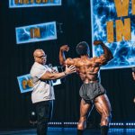 Hosts - Back view of a Shirtless Man Flexing His Muscles while on Stage